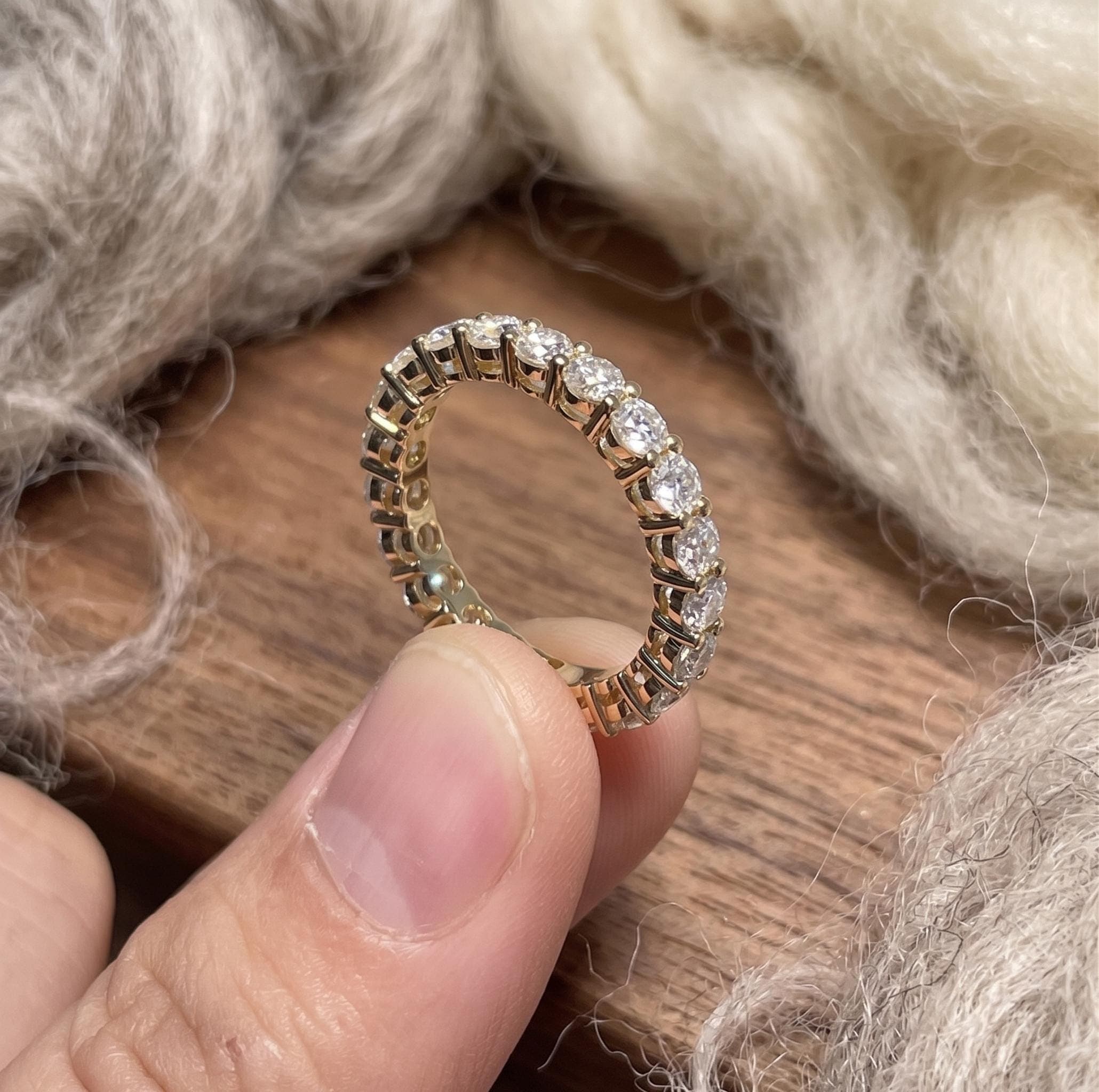 May include: A gold eternity ring featuring a continuous line of round, clear gemstones. The ring is held in a hand, highlighting the detailed setting and sparkle of the stones. The ring is set against a neutral backdrop.